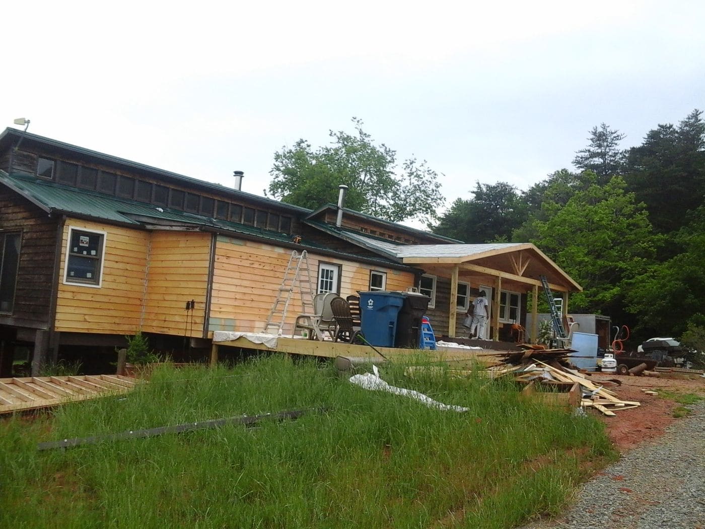 Wooden residential cabin under construction with green metal roof, ladders, and building materials visible in yard surrounded by trees